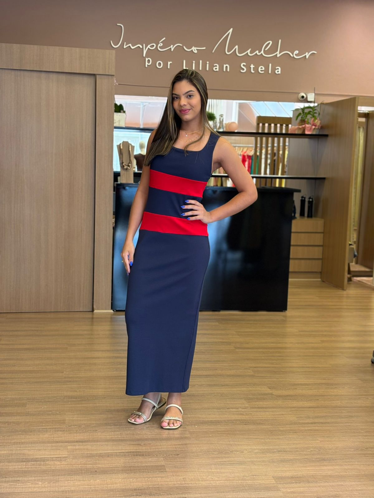 Modelo vestindo vestido longo azul marinho com faixas horizontais vermelhas, sem mangas, modelagem reta e elegante.