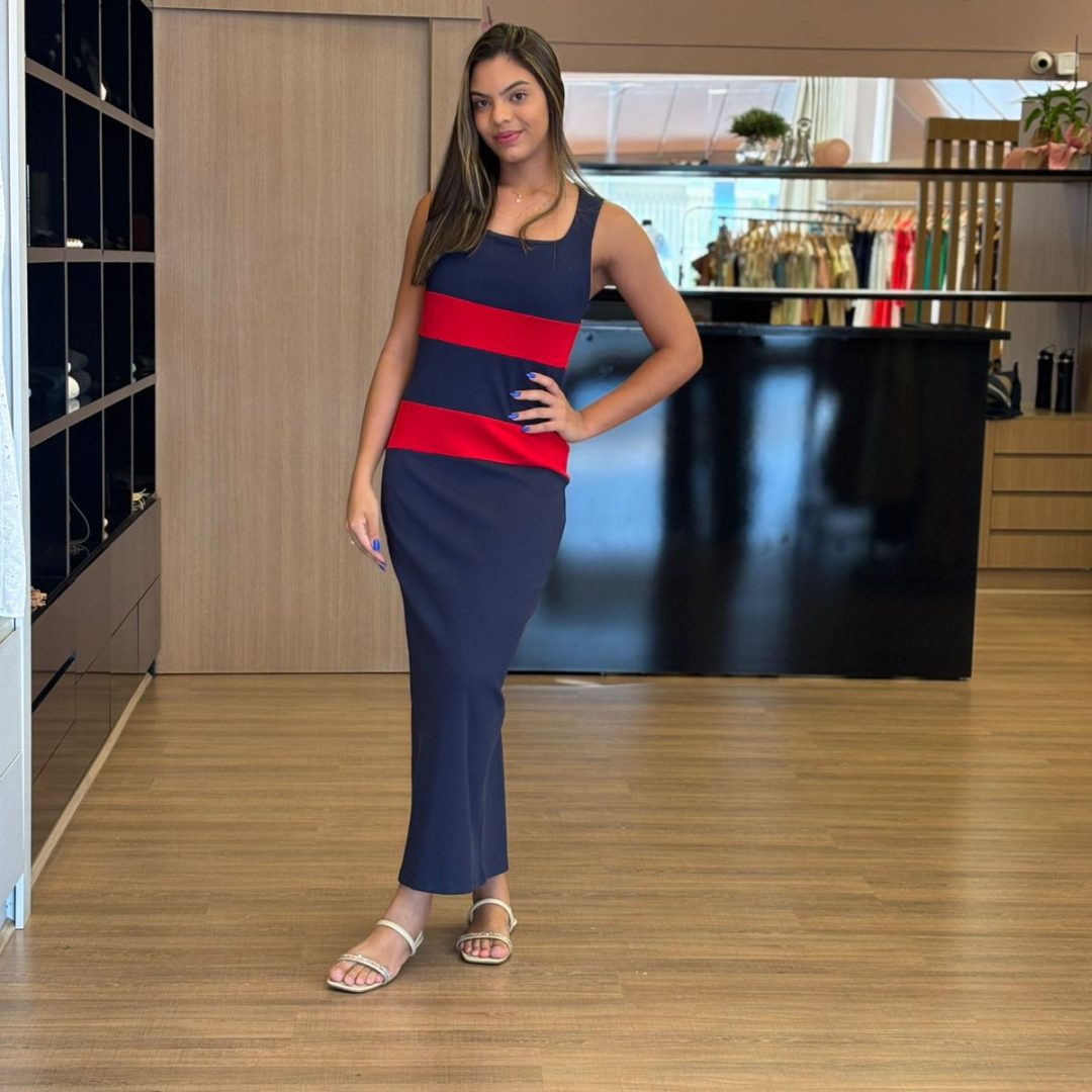 Modelo vestindo vestido longo azul marinho com faixas horizontais vermelhas, sem mangas, modelagem reta e elegante.
