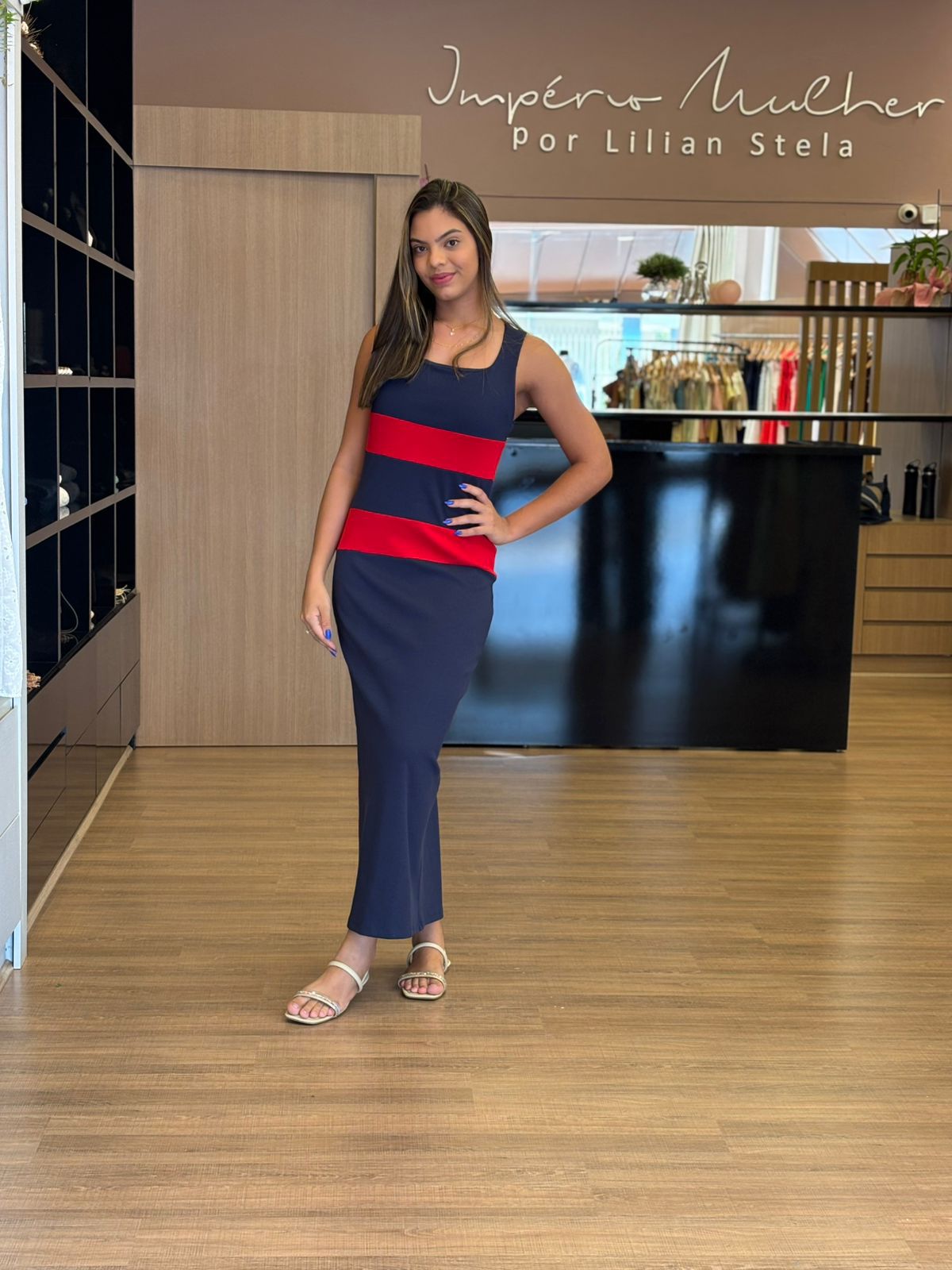 Modelo vestindo vestido longo azul marinho com faixas horizontais vermelhas, sem mangas, modelagem reta e elegante.
