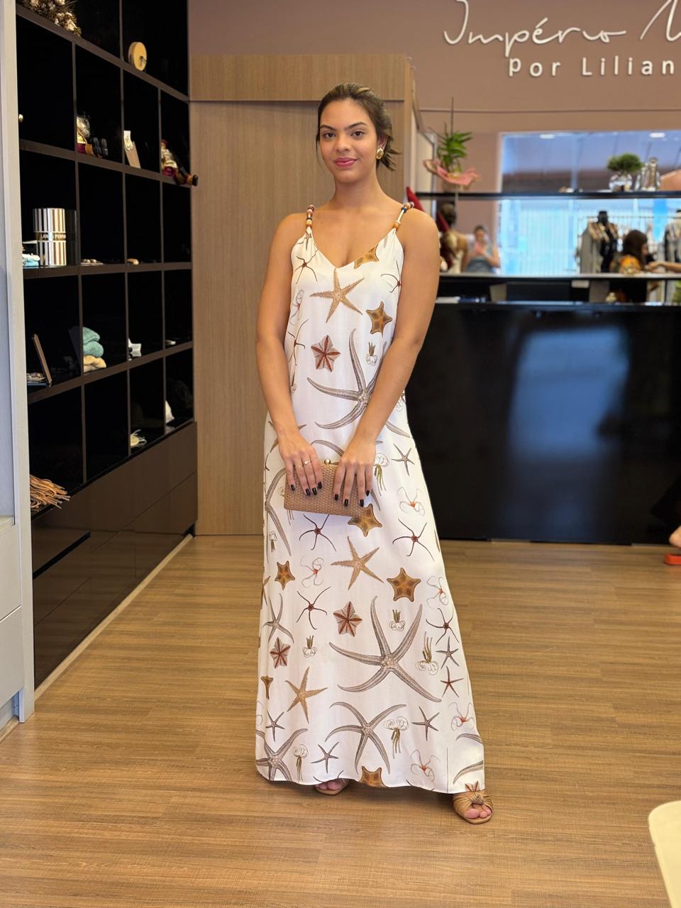 Modelo usando vestido longo branco com estampa marinha em tons neutros, alças finas com detalhes, modelagem fluida e caimento leve, em ambiente interno de loja.