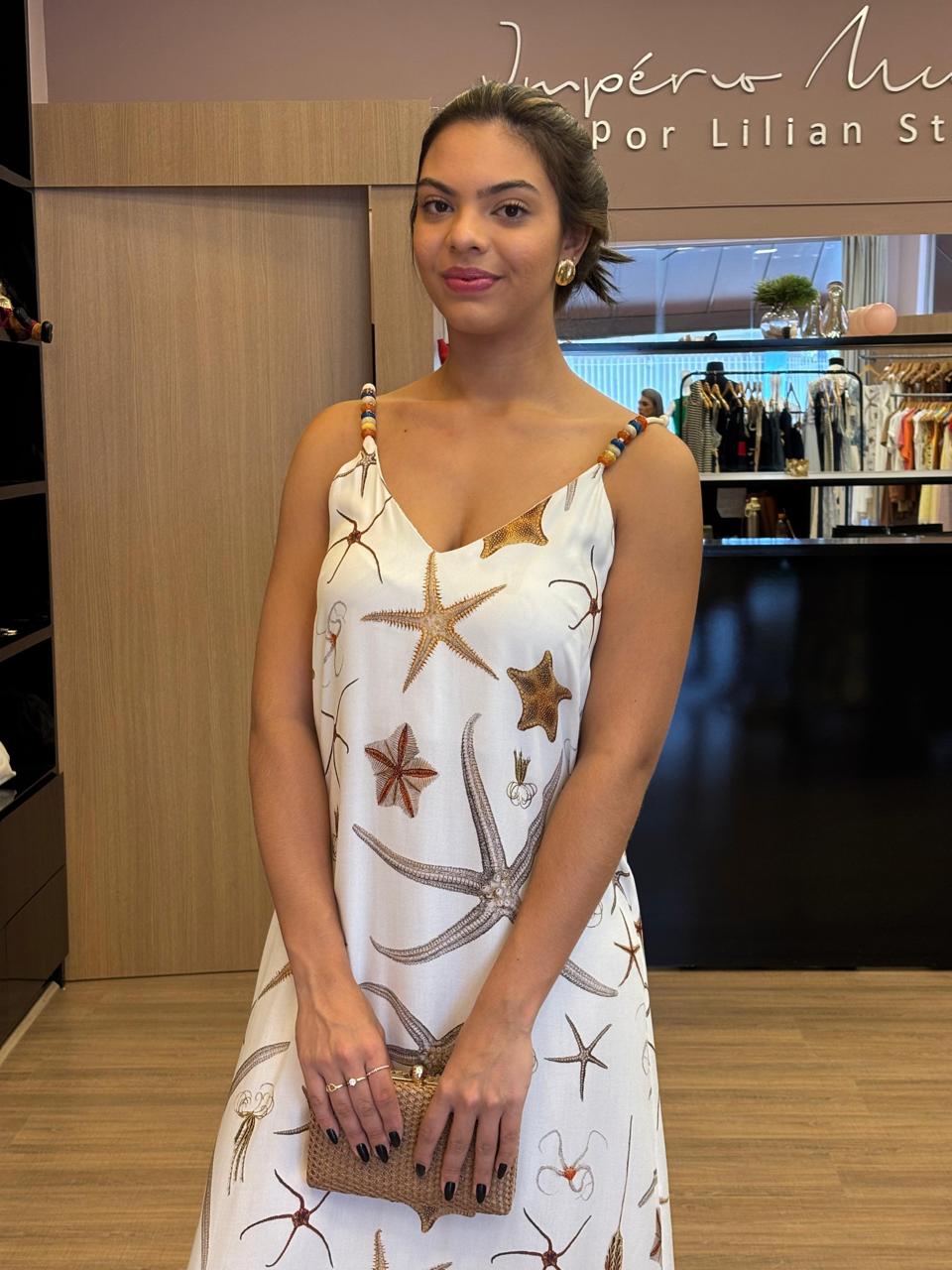 Modelo usando vestido longo branco com estampa marinha em tons neutros, alças finas com detalhes, modelagem fluida e caimento leve, em ambiente interno de loja.