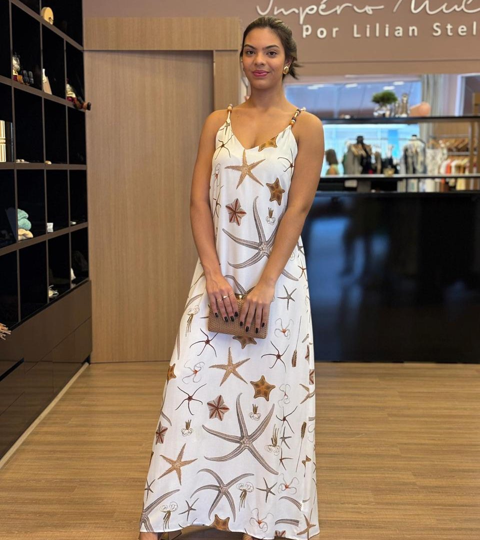 Modelo usando vestido longo branco com estampa marinha em tons neutros, alças finas com detalhes, modelagem fluida e caimento leve, em ambiente interno de loja.