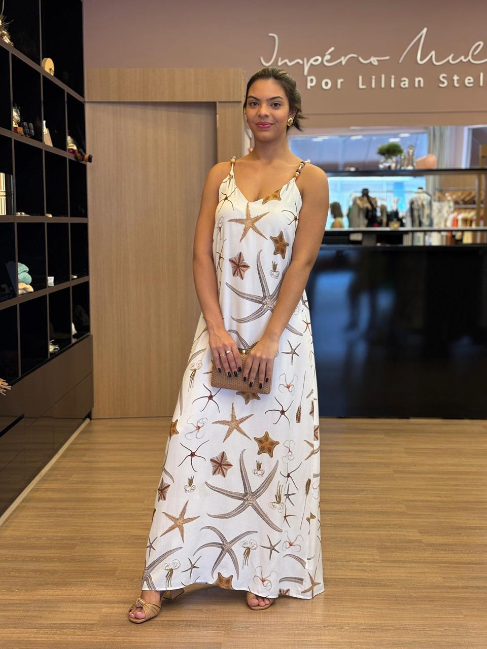 Modelo usando vestido longo branco com estampa marinha em tons neutros, alças finas com detalhes, modelagem fluida e caimento leve, em ambiente interno de loja.