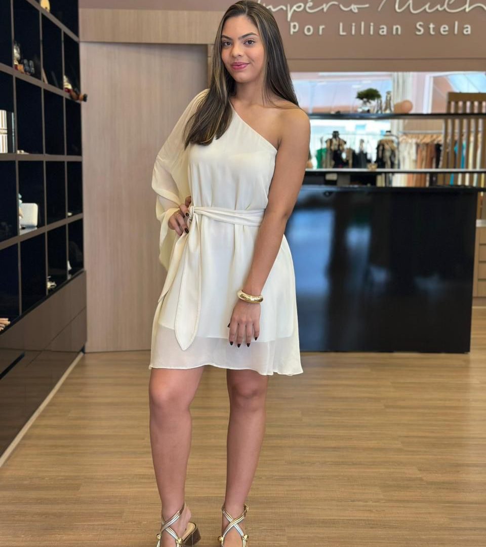 Modelo usando vestido curto claro de um ombro só, com manga fluida em um lado, faixa para amarração na cintura e modelagem leve, em ambiente interno de loja.