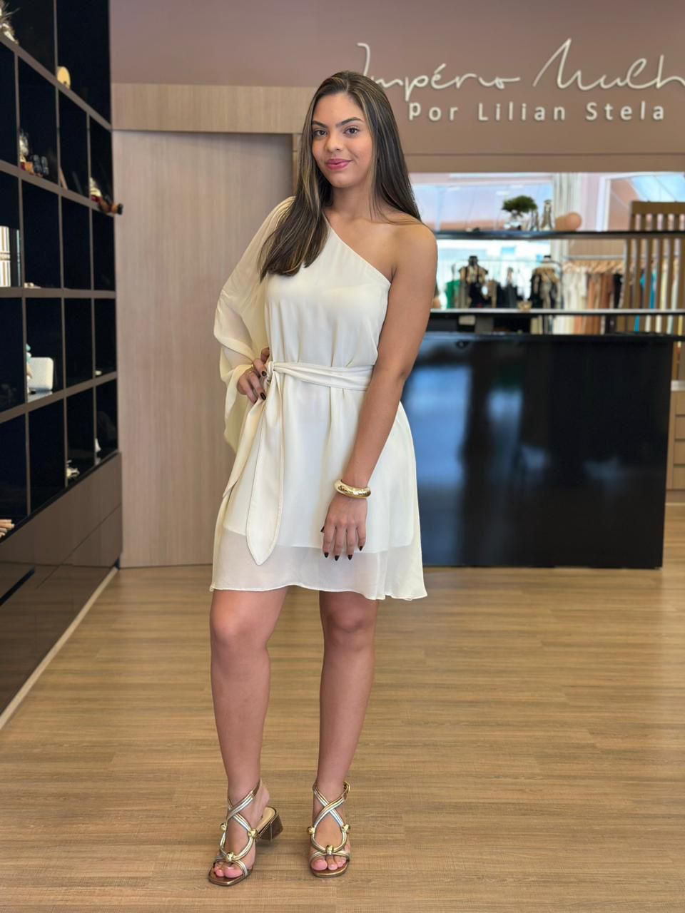 Modelo usando vestido curto claro de um ombro só, com manga fluida em um lado, faixa para amarração na cintura e modelagem leve, em ambiente interno de loja.
