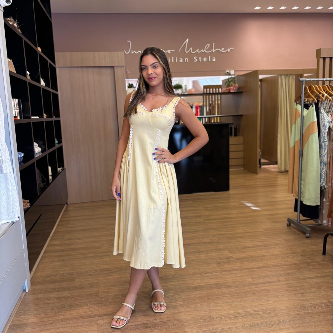 Modelo usando vestido midi amarelo claro, com alças médias, detalhes em renda branca no busto e ao longo da peça, modelagem acinturada e saia fluida.