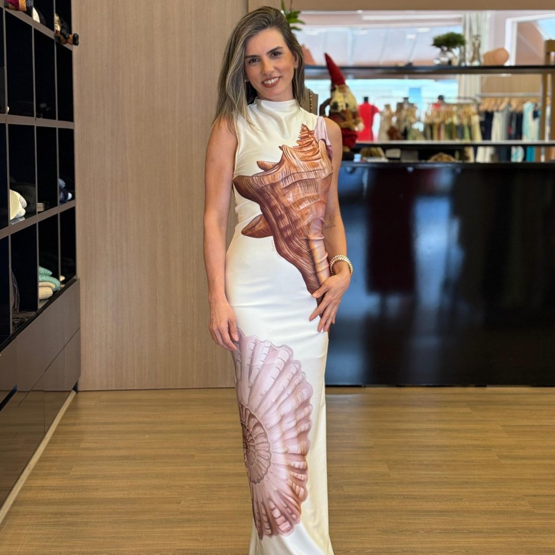 Mulher usando vestido midi off white, sem mangas, com estampa grande em tons terrosos inspirada em formas naturais, modelagem ajustada ao corpo e caimento elegante, em ambiente interno de loja.