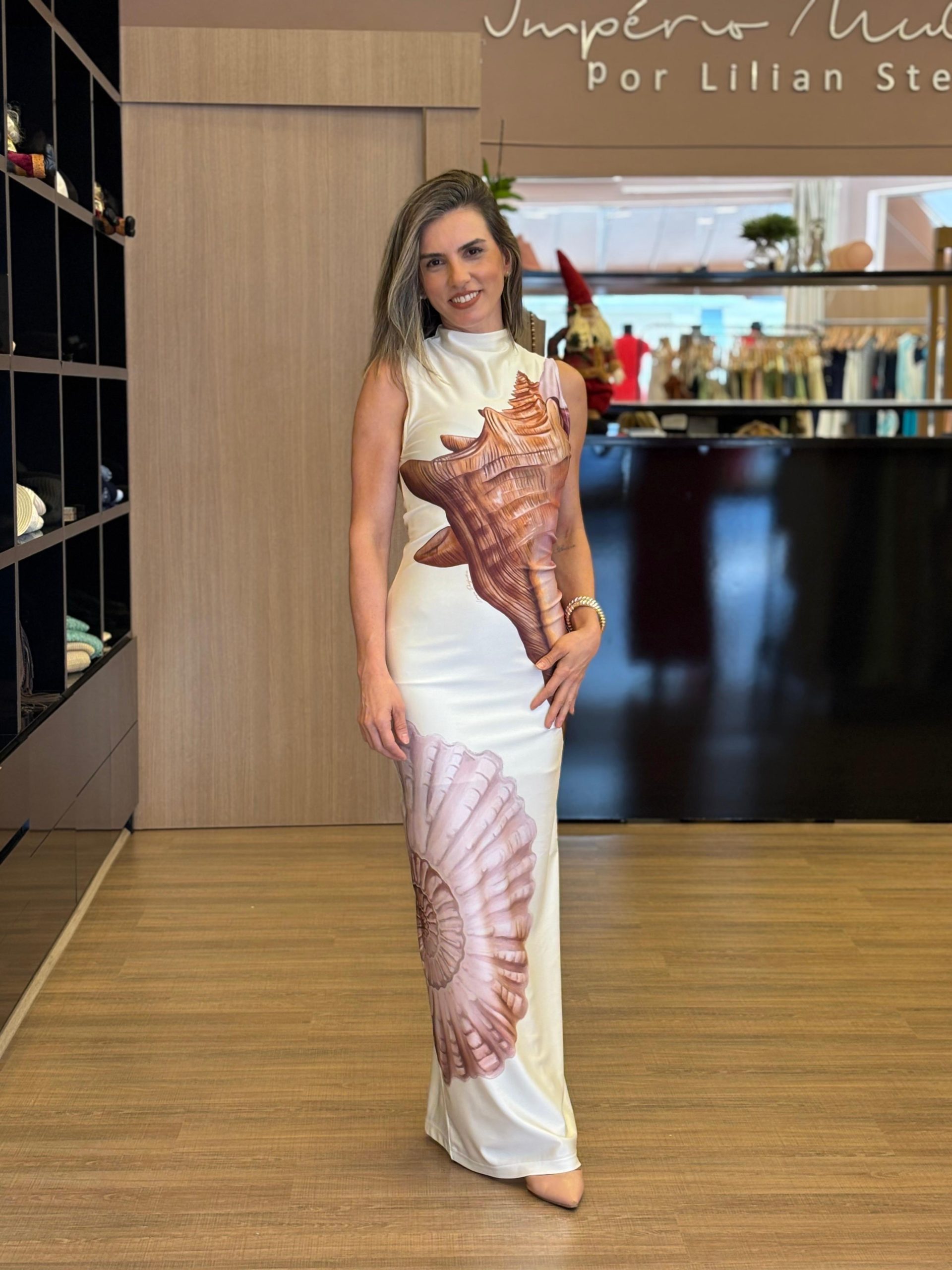 Mulher usando vestido midi off white, sem mangas, com estampa grande em tons terrosos inspirada em formas naturais, modelagem ajustada ao corpo e caimento elegante, em ambiente interno de loja.