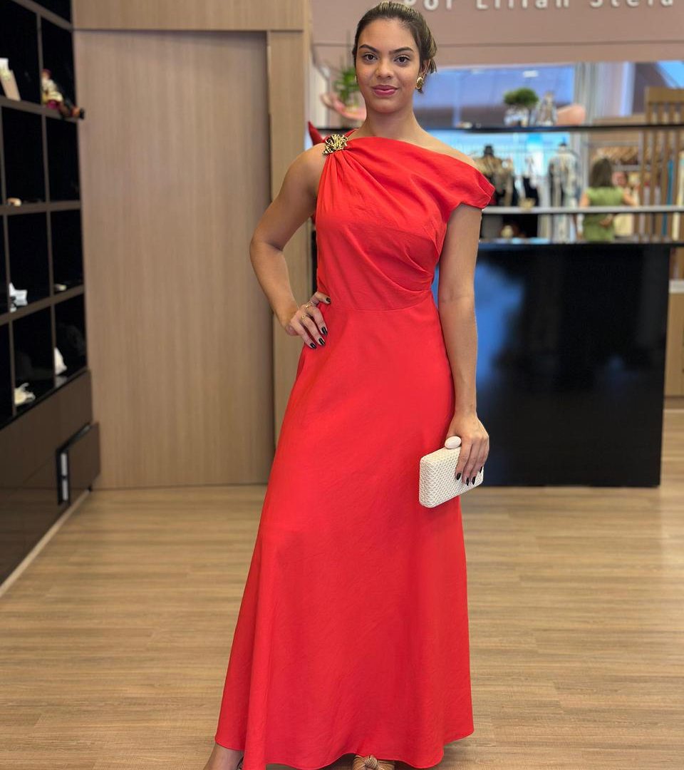 Modelo vestindo vestido longo vermelho de um ombro só, com detalhe de broche dourado no ombro.