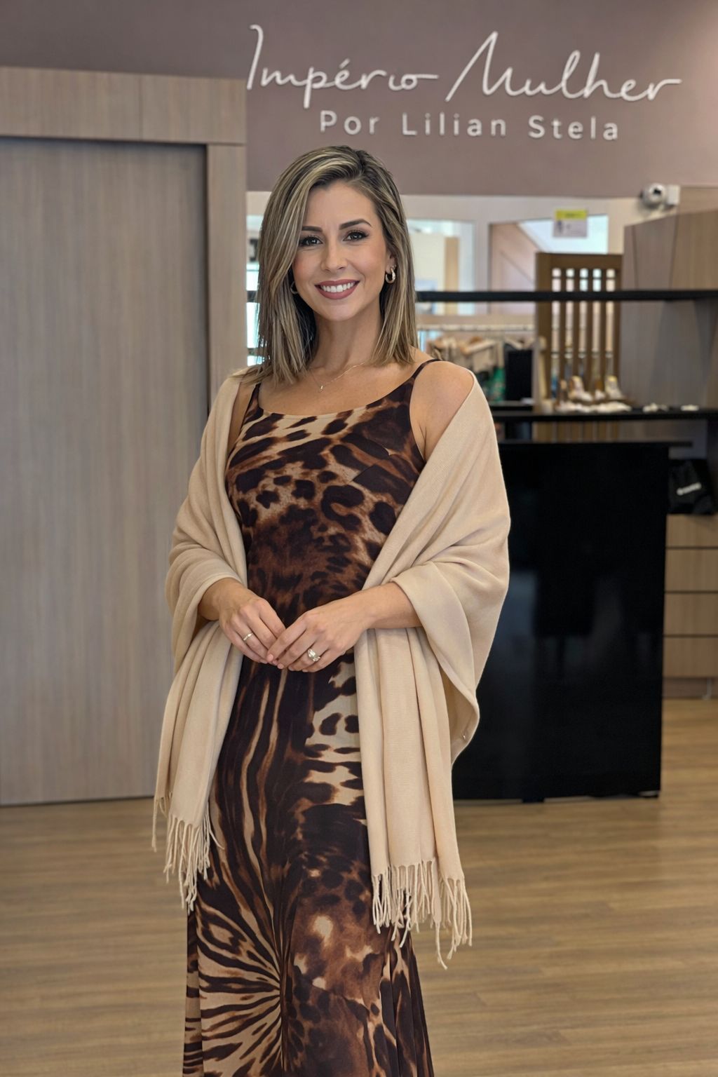 Modelo usando pashmina feminina com franjas sobre vestido estampado marrom, em ambiente interno de loja, compondo um look elegante e sofisticado.