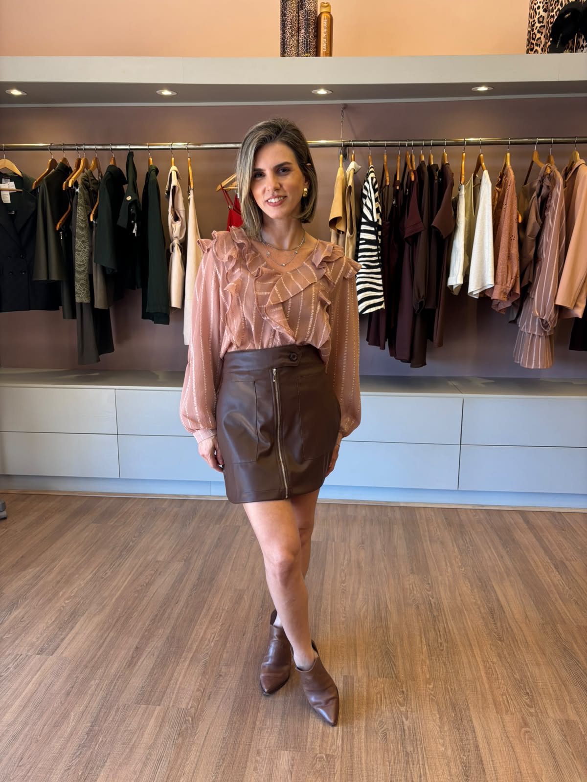 Saia curta marrom em couro ecológico com zíper frontal, combinada com blusa estampada de mangas longas e botas de bico fino.
