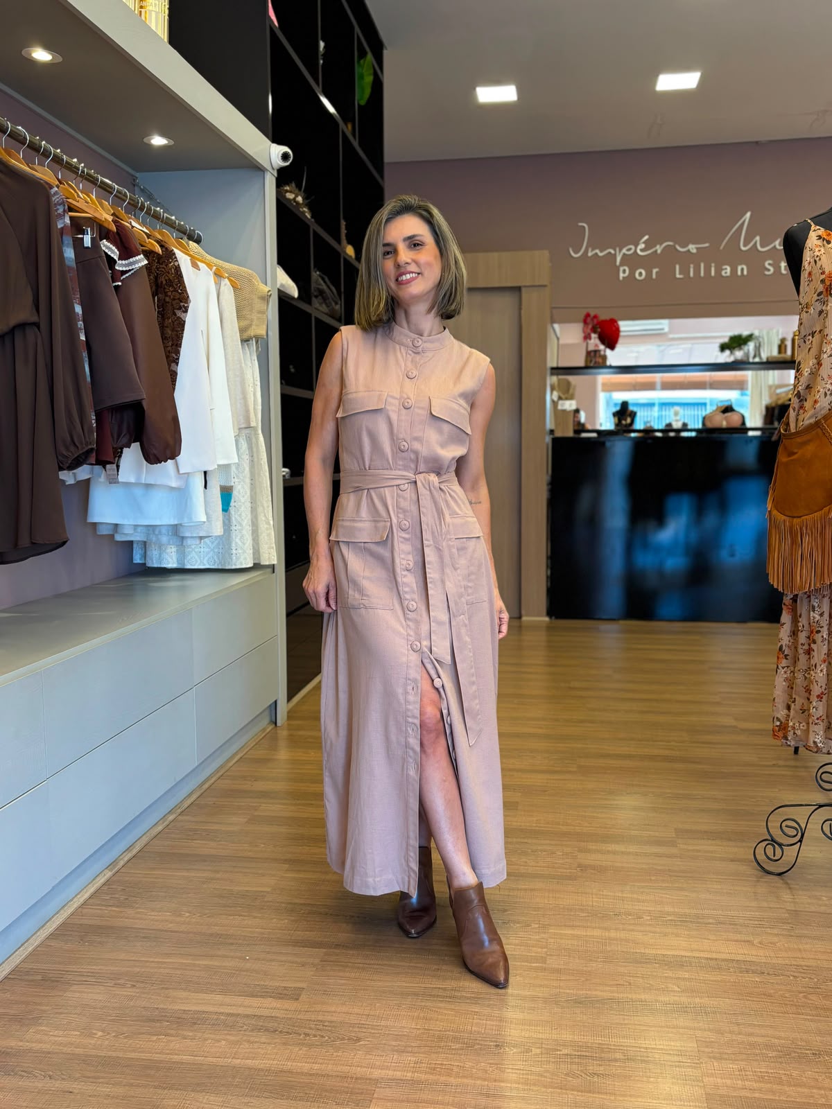 Modelo vestindo vestido chemise longo bege sem mangas, com botões frontais, bolsos e faixa para amarração na cintura, em ambiente de loja feminina.