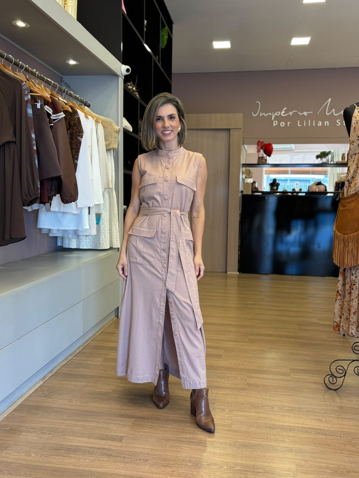Modelo vestindo vestido chemise longo bege sem mangas, com botões frontais, bolsos e faixa para amarração na cintura, em ambiente de loja feminina.