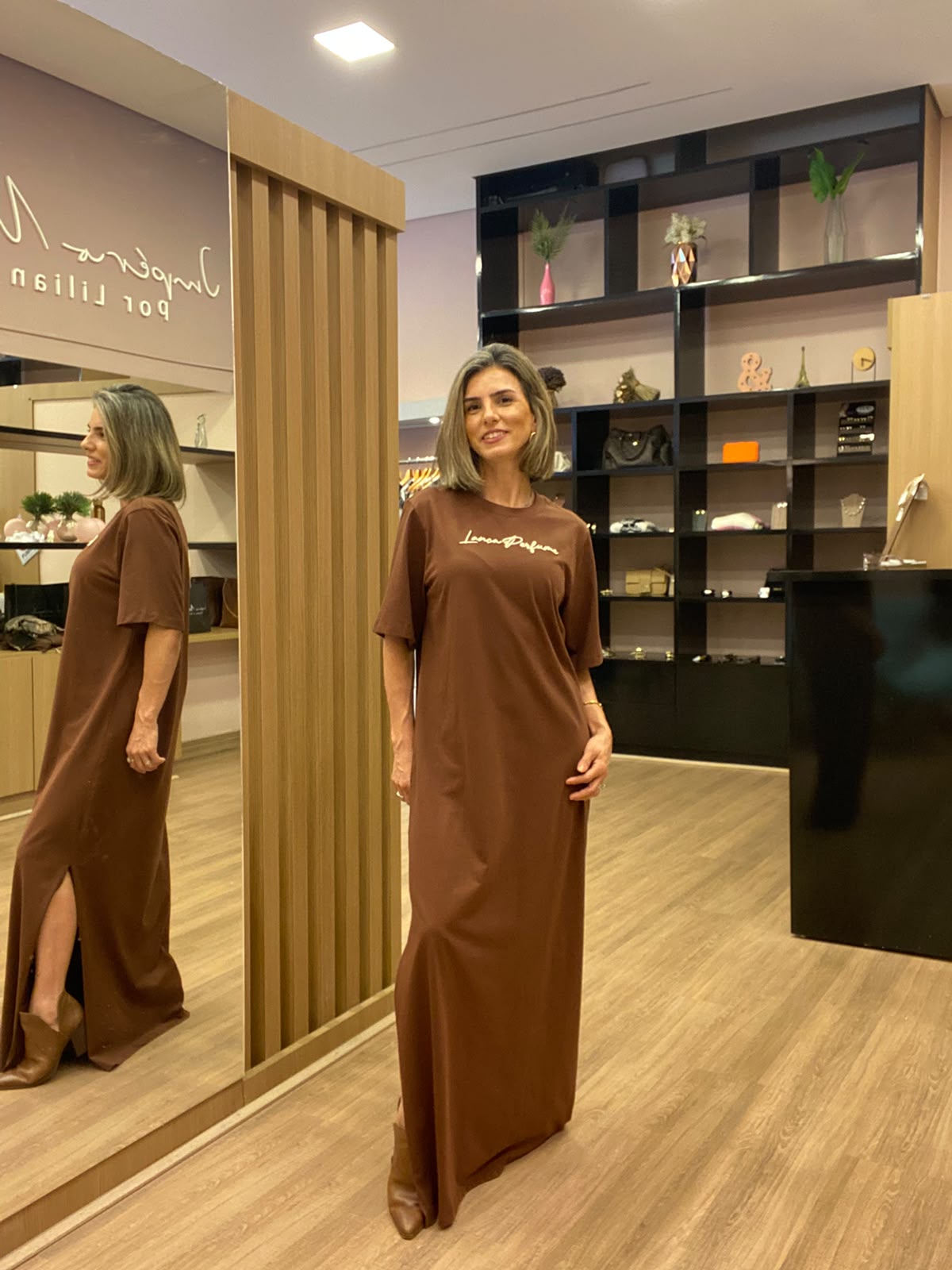 Modelo usando vestido longo marrom com mangas curtas, detalhe drapeado na cintura e fenda lateral, em ambiente de loja feminina.