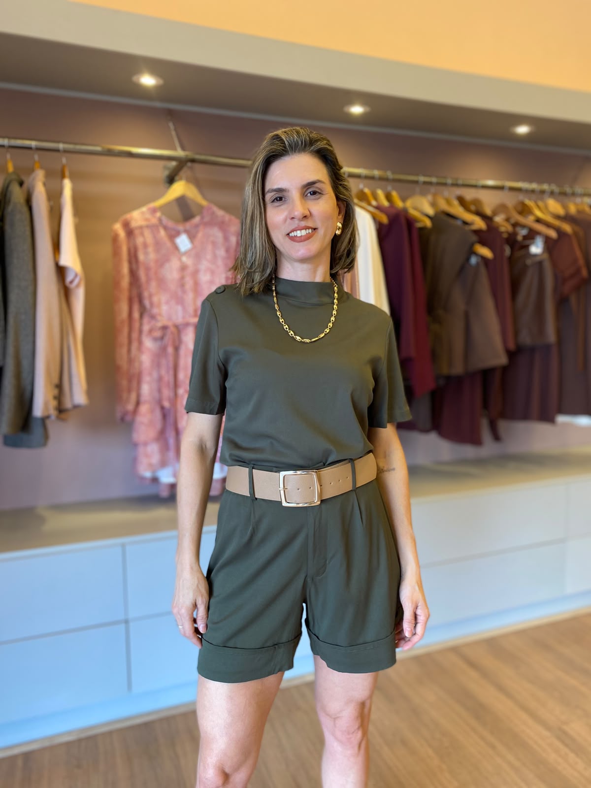 Modelo veste conjunto feminino verde militar com blusa de manga curta, short de alfaiataria com pregas e cinto largo bege.