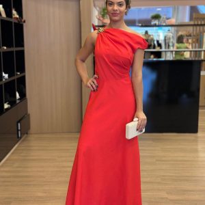 Modelo vestindo vestido longo vermelho de um ombro só, com detalhe de broche dourado no ombro.