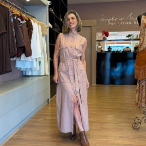 Modelo vestindo vestido chemise longo bege sem mangas, com botões frontais, bolsos e faixa para amarração na cintura, em ambiente de loja feminina.