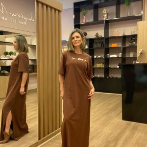 Modelo usando vestido longo marrom com mangas curtas, detalhe drapeado na cintura e fenda lateral, em ambiente de loja feminina.